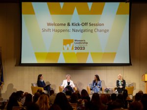 Four women are seated on chairs on a stage. The screen overhead reads: Welcome and Kick-Off Session, Shift Happens: Navigating Change, Women's Leadership Conference 2023. The sign on the podium reads: ӰƬ University.