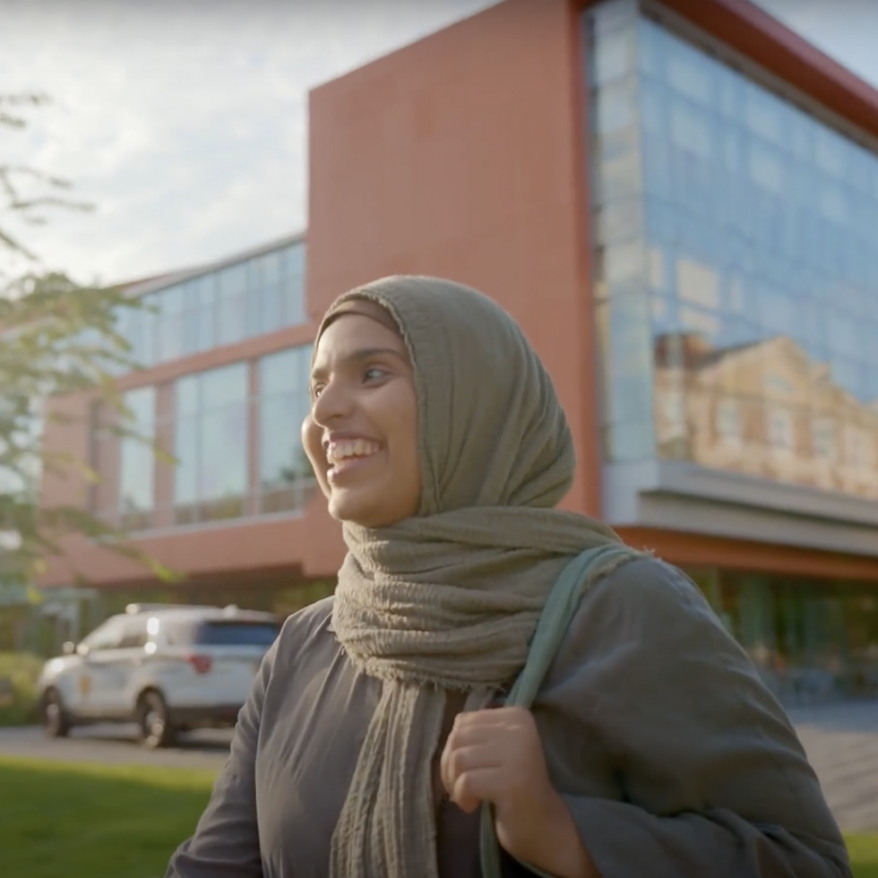 An ӰƬ student smiling and walking in front of the Nexus building.