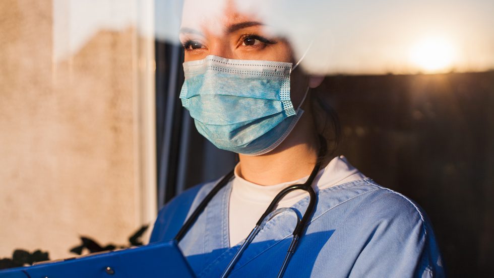 Young sad female caucasian UK US GP EMS doctor carer looking through ICU window,fear uncertainty in eyes,wearing face mask gazing at sun,hope faith in overcoming Coronavirus COVID-19 pandemic crisis