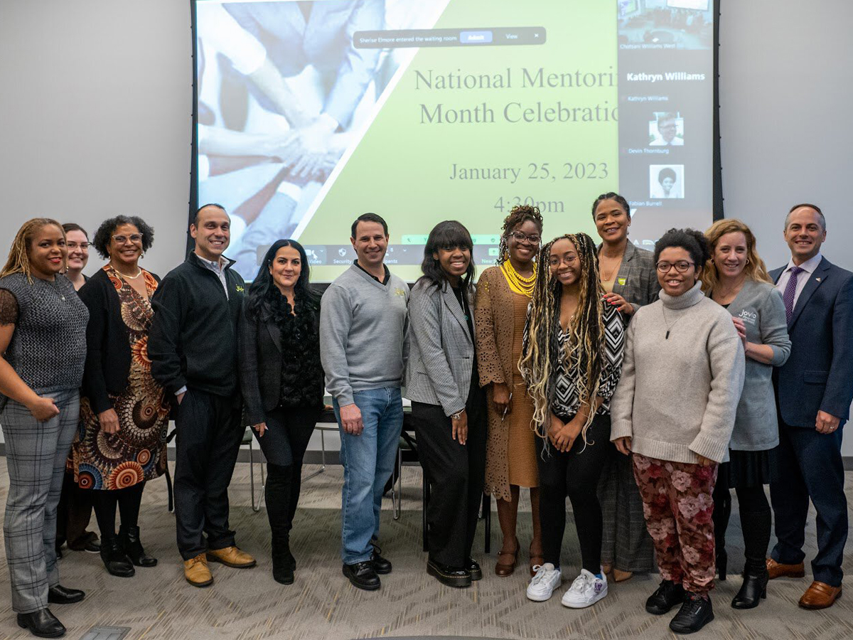 Students and mentors are standing in front of a screen that reads "National Mentoring Month Celebration, January 25, 2023, 4:30 p.m." To the left is a microphone at a podium with the words "ӰƬ University