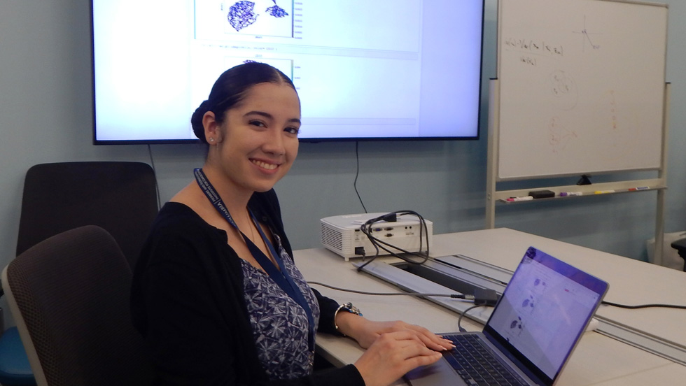 A woman with dark hair in a bun, smiling, wearing a black-and-white print shirt, sits in front of a laptop. On the screen behind her are scientific illustrations.