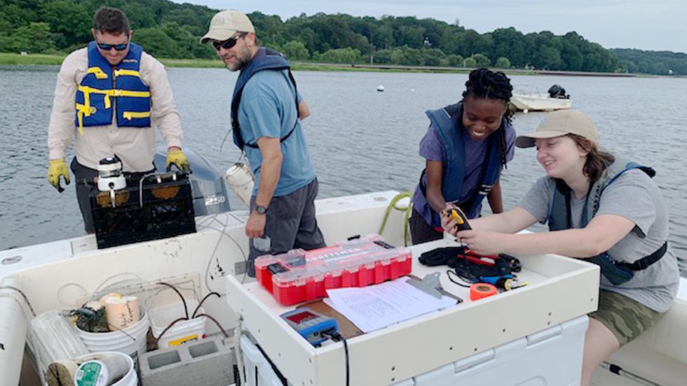 A photograph of ӰƬ faculty members Ryan Wallace and Aaren Freeman aboard a boat filled with scientific equipment off the Long Island shoreline. Two student assistants are on the boat as well.