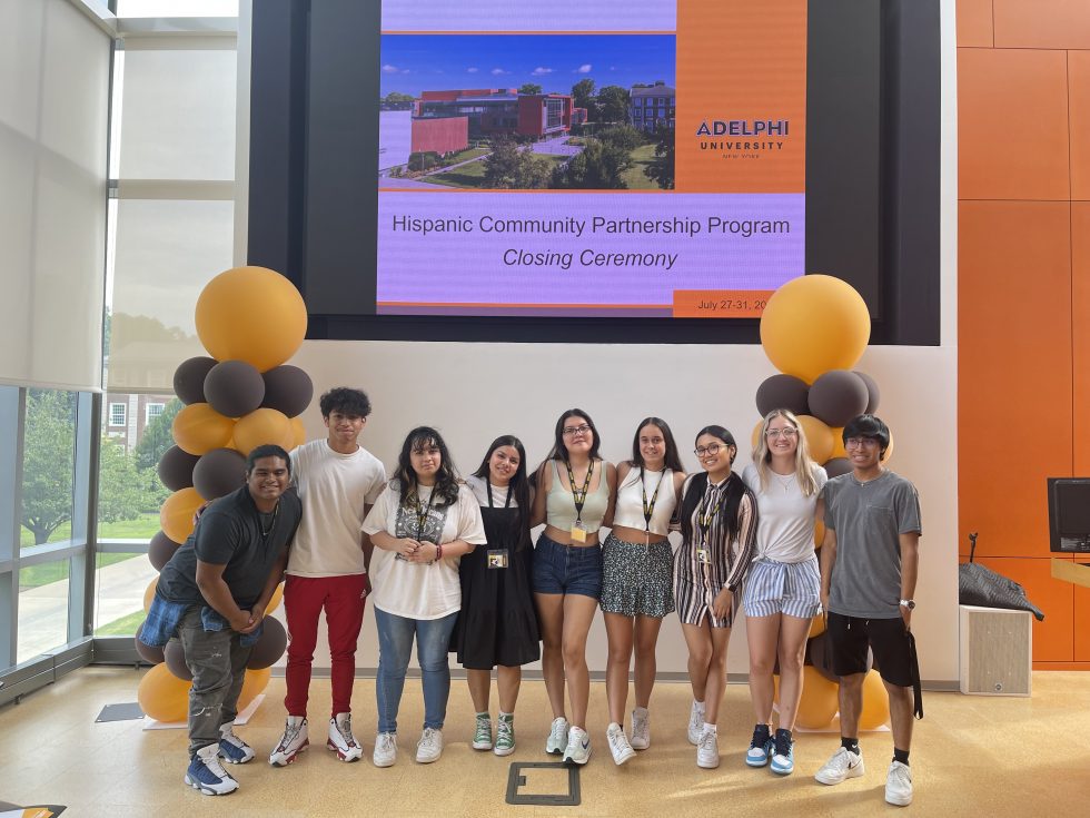 Nine students, male and female, of Hispanic descent, stand in front of a screen with the words “Hispanic Community Partnership Program Closing Ceremony: ӰƬ University” and a photo of the ӰƬ University campus. To their left and right are brown and gold balloons.