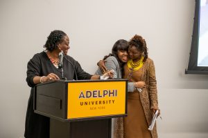Black woman with brown hair and gold earrings wearing a brown dress stands at a podium with the words ӰƬ University. A young Black woman wearing a gray blazer is on her left, smiling and hugging a Black woman with braids, also smiling, wearing a brown jacket with a yellow collar and brown skirt.