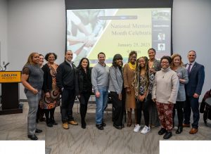 Thirteen people, male and female, of various ages and backgrounds, stand in front of a screen that reads National Mentoring Month Celebration: ӰƬ University.