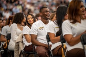 A young Black man is sitting in an audience with people seated in front of and behind him. They wear ӰƬ University College of Nursing and Public Health scrubs.