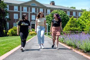 Three white young women walk on a path in front of ӰƬ’s Levermore Hall. There are purple flowers growing on the right.