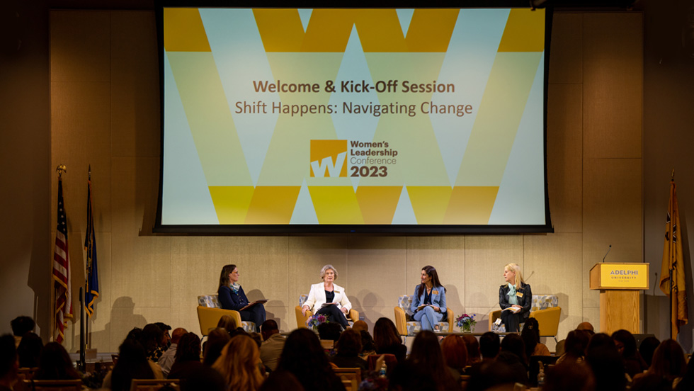 Four women are seated on chairs on a stage. The screen overhead reads: Welcome and Kick-Off Session, Shift Happens: Navigating Change, Women's Leadership Conference 2023. The sign on the podium reads: ӰƬ University.