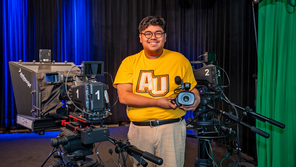 A young man in glasses and a gold shirt that reads AU鈥攆or 成人影片 University鈥� is smiling, holding a camera. On the left is a camera labeled 1 Autoscript, and on the right, a camera labeled 2.