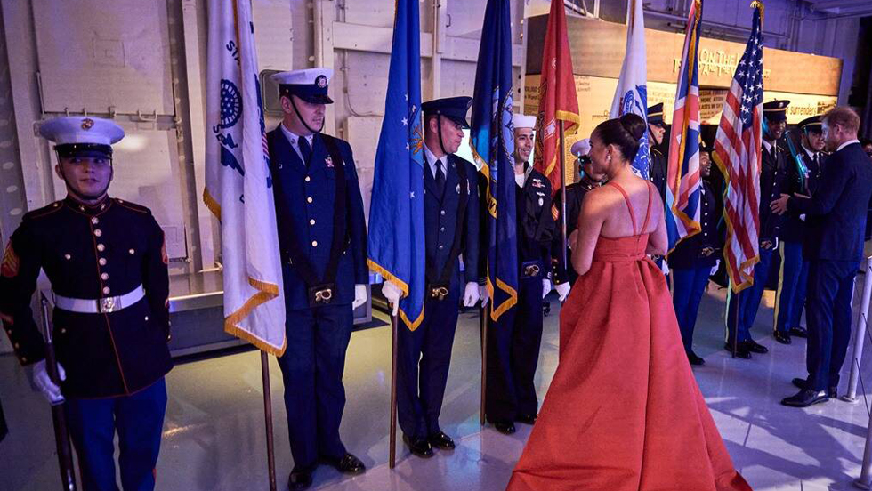 A row of military servicemen in full-dress uniform holding flags at a gala, greeting a woman wearing a formal, full-length red gown, whose back is to the camera