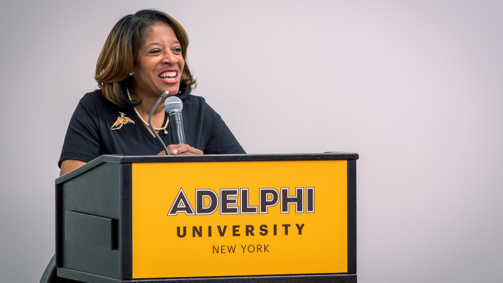 Linda Davis Valdez smiles broadly while giving her address. She is standing before a microphone on a lectern with a gold sign saying 成人影片 University, New York.