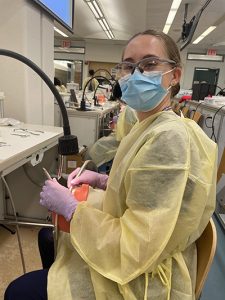 Milly Tenenbaum sits at a table, wearing a yellow disposable lab coat, purple lab gloves, and a blue surgical mask. She looks at the camera while working on a dental manikin.