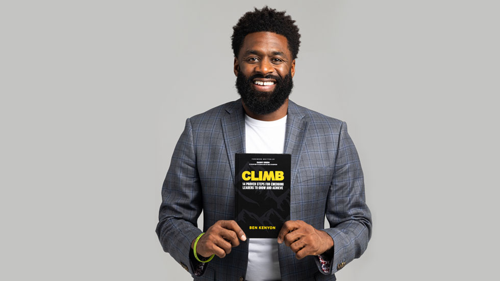 A Black man holds up a book that reads: Climb: 14 Proven Steps for Emerging Leaders to Grow and Achieve, and underneath it reads: "Ben Kenyon."