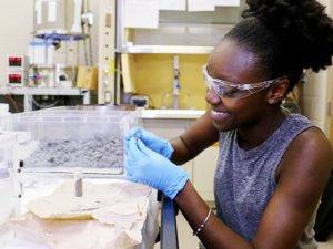 Keanna Jardine wears protective glasses and gloves as she handles a gray substance similar to moon rocks.