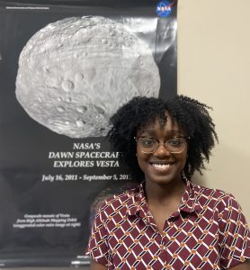 Dr. Jardine stands in front of a poster that says "NASA's Dawn Spacecraft Explores Vesta: July 16, 2011鈥� September 5, 2012." The poster includes a large picture of the asteroid Vesta, which is the second most massive object in the asteroid belt.
