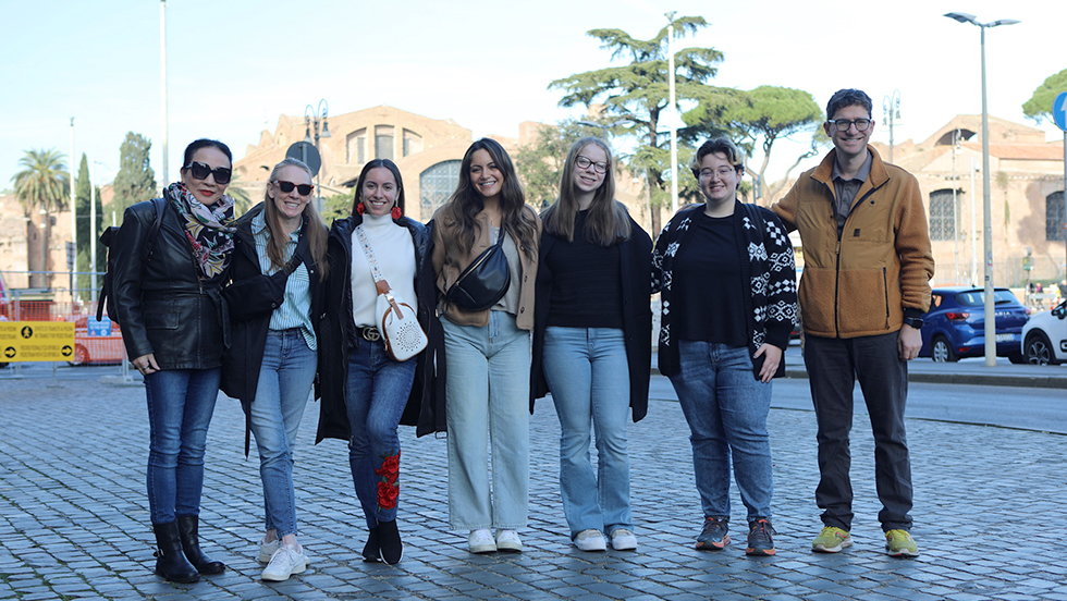 uring a recent trip to Italy, an adult female (left) with short black hair, sunglasses, a short leather jacket, multicolored scarf. jeans and boots stands with six students—also in casual wear—five females and one male (right).Optional: They are standing in a plaza with gray pavers; two beige buildings and trees are in the background.