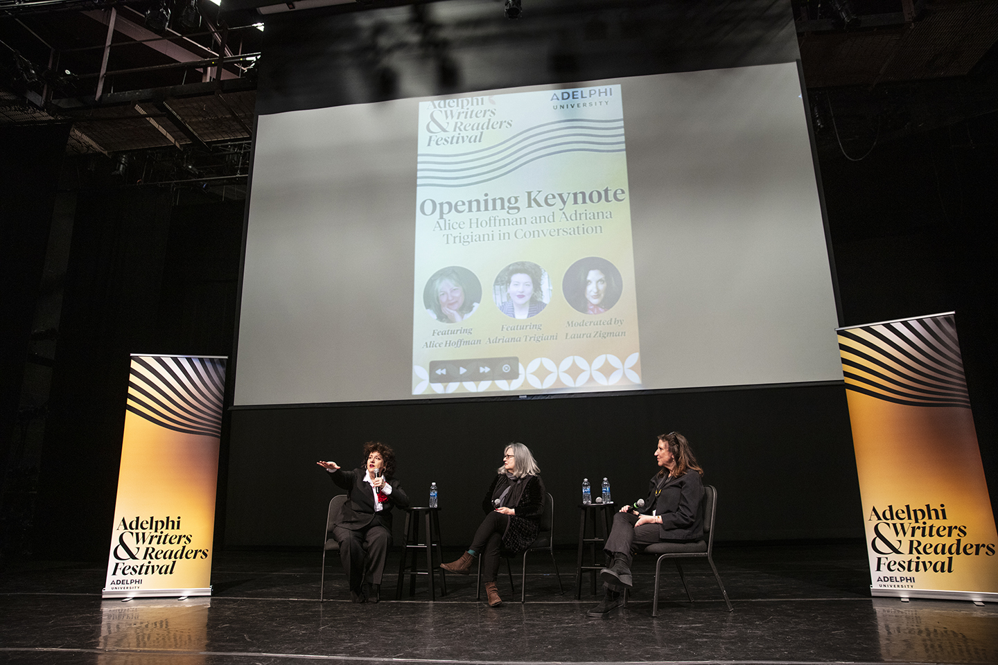 Authors and moderator seated on stage. There are banners on each side of them that read “ӰƬ Writers & Readers Festival” and a screen above them that says “Opening Keynote: Alice Hoffman and Adriana Trigiani in Conversation.” The screen also has their photos, along with a photo of the moderator.
